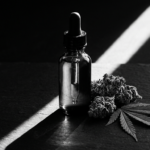 Fotografia de alto contraste em preto e branco de um frasco conta-gotas de vidro, botões de flor de cannabis e uma folha de cannabis sobre uma superfície de pedra escura, com um feixe de luz afiado cortando a cena e criando sombras dramáticas.
